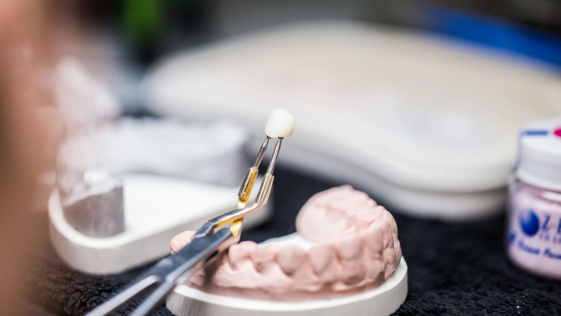Tweezers holding a ceramic crown above a pink dental model in a dental laboratory