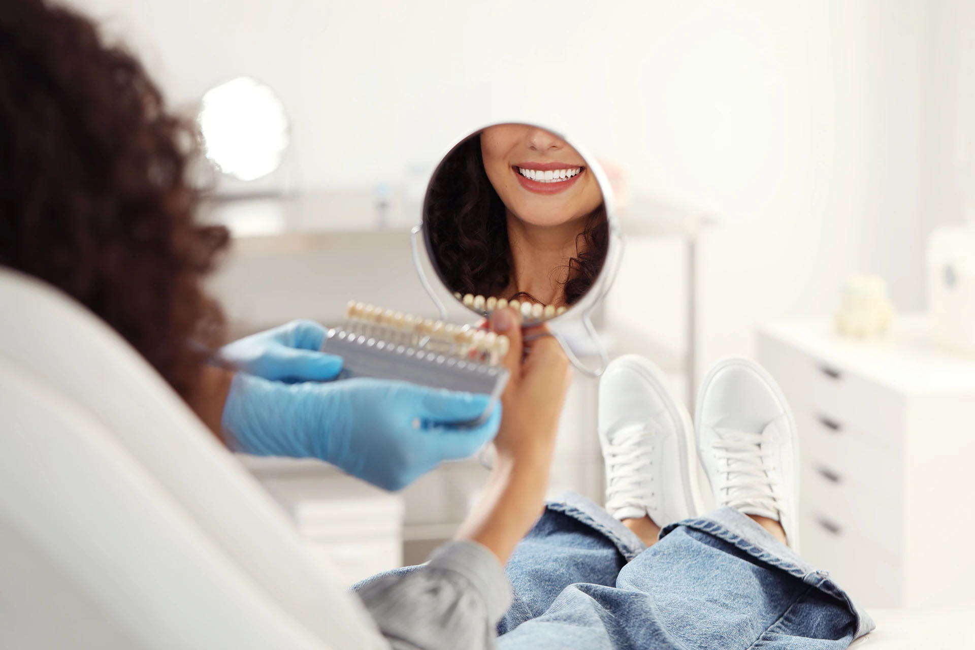 Woman in a dental chair smiling into a hand mirror as a dentist holds a shade guide
