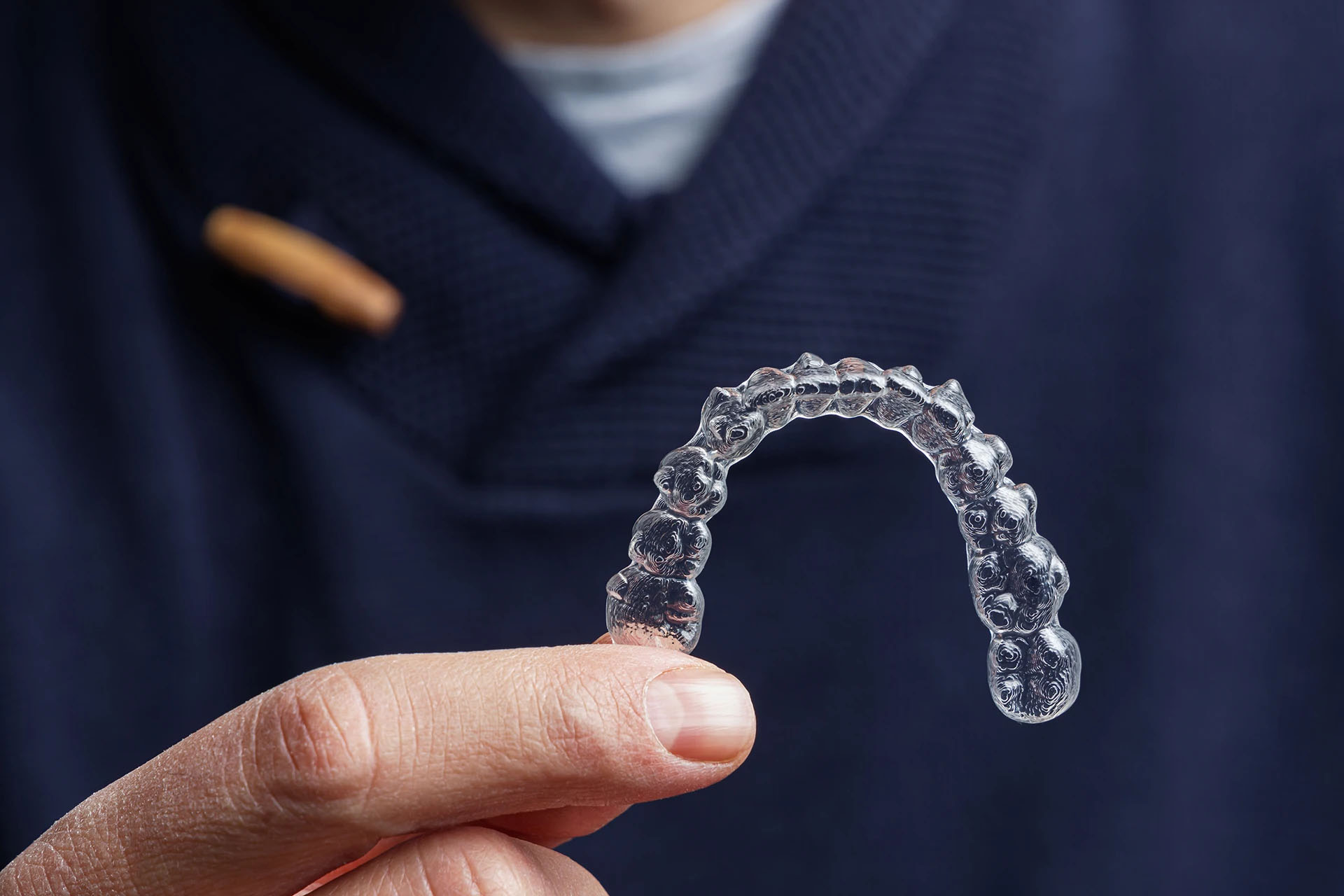 A person in a navy sweater holding a transparent clear aligner between their fingers