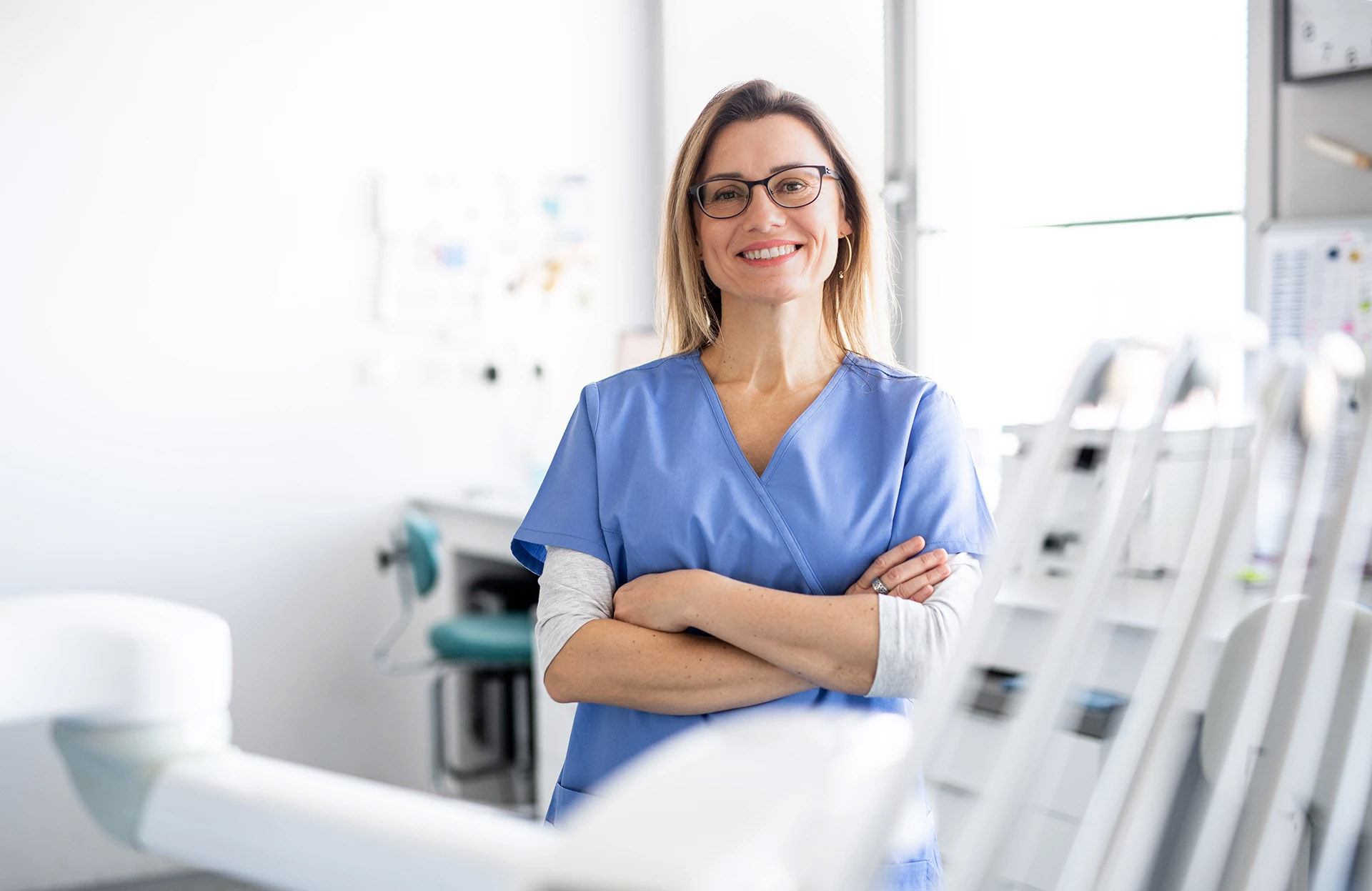 Smiling female dentist in blue scrubs and glasses standing with arms crossed in the treatment room