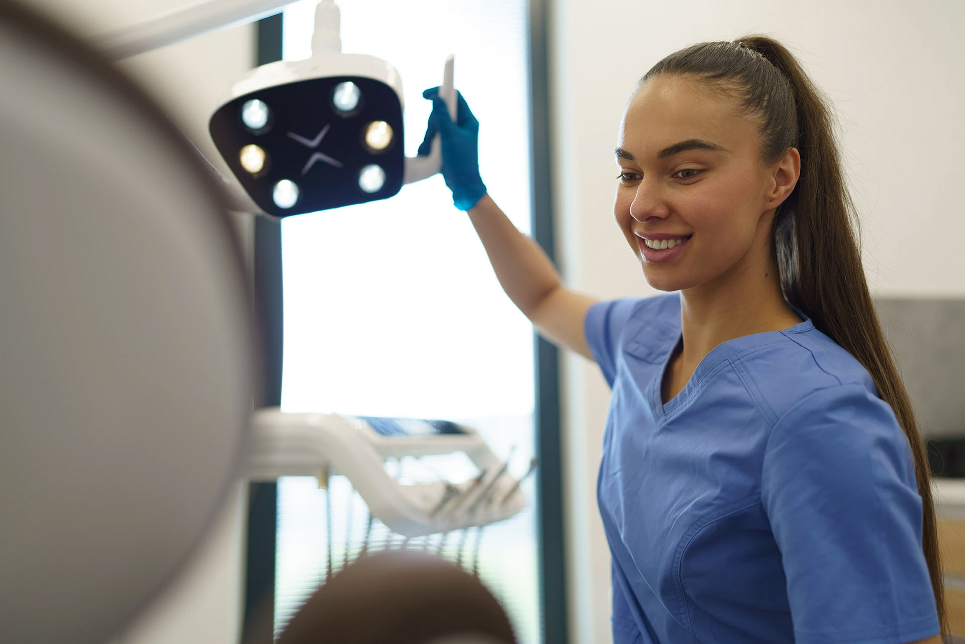 Smiling dental assistant in blue scrubs adjusting the overhead operatory light