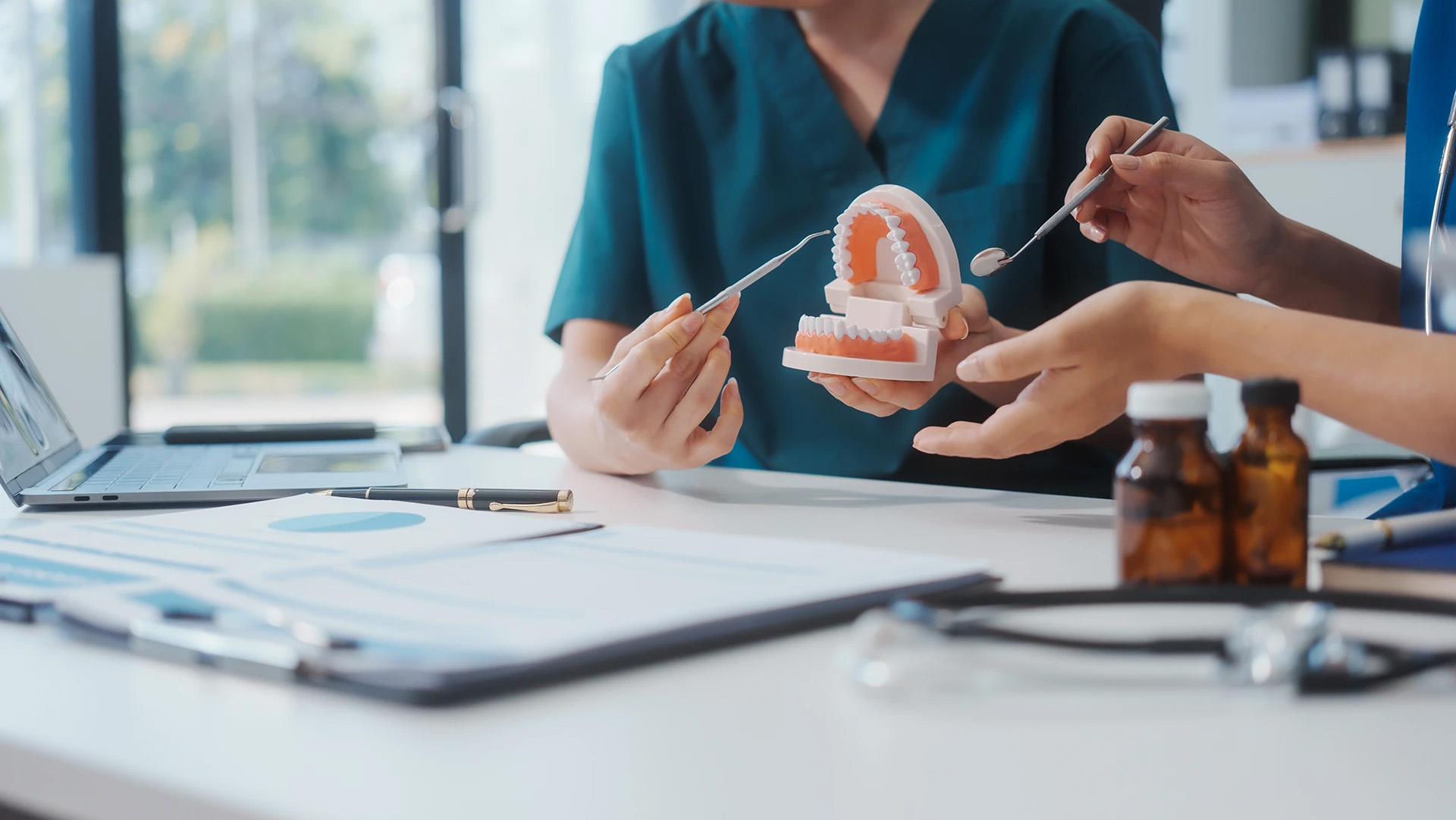 Two dental professionals examining a teeth model with instruments at a desk with charts and medication bottles