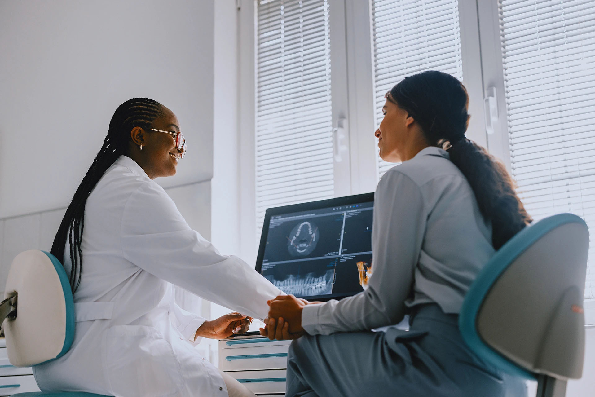 Dentist in a white coat holding a patient's hands and smiling during a consultation with a dental scan on the monitor