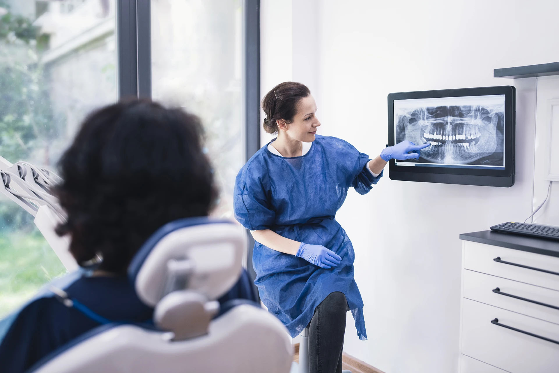 Dentist in a blue gown pointing to a panoramic dental X-ray on a wall monitor for a seated patient