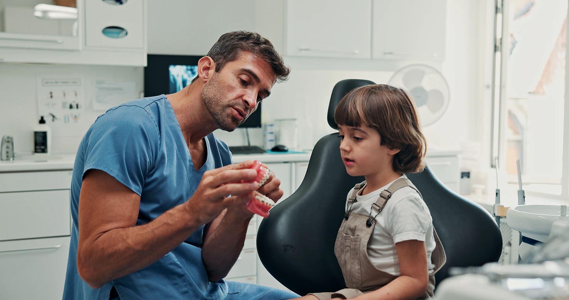 Male dentist in blue scrubs showing a teeth model to an attentive young boy in the dental chair