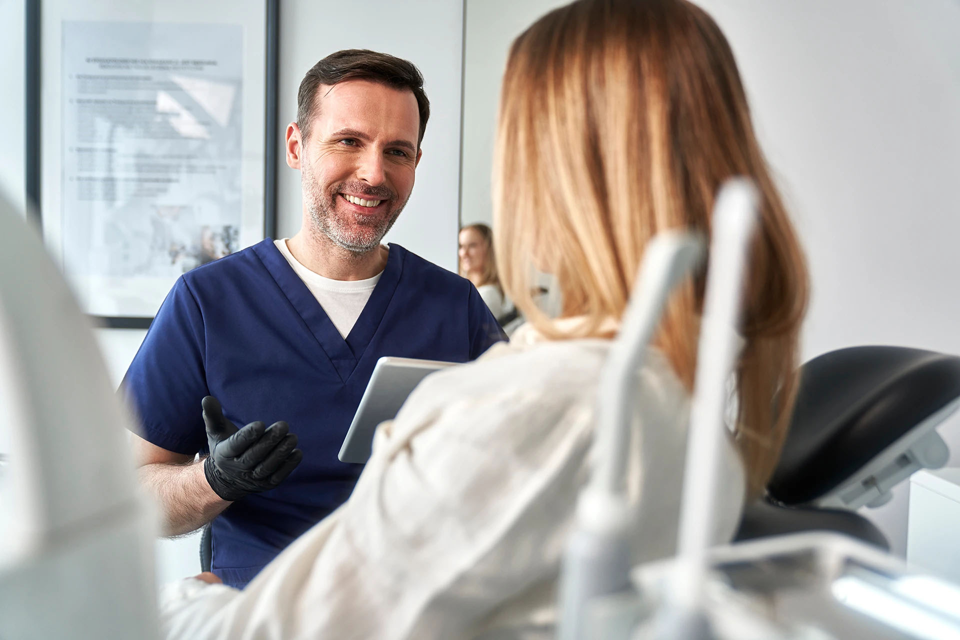 Smiling male dentist in navy scrubs holding a tablet while talking with a patient in the chair