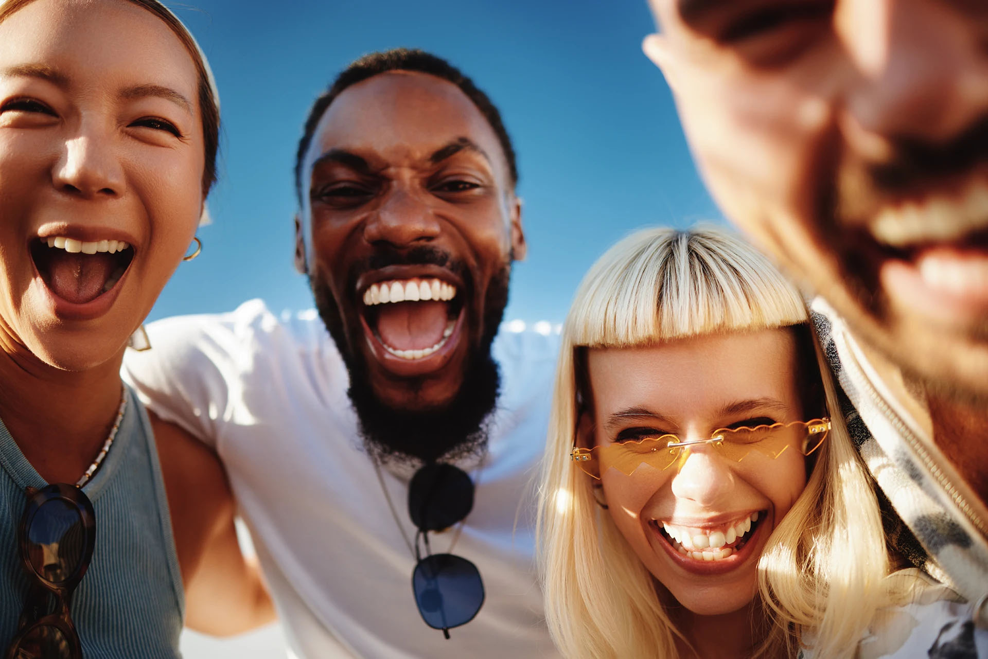 Four diverse friends laughing together outdoors under a blue sky