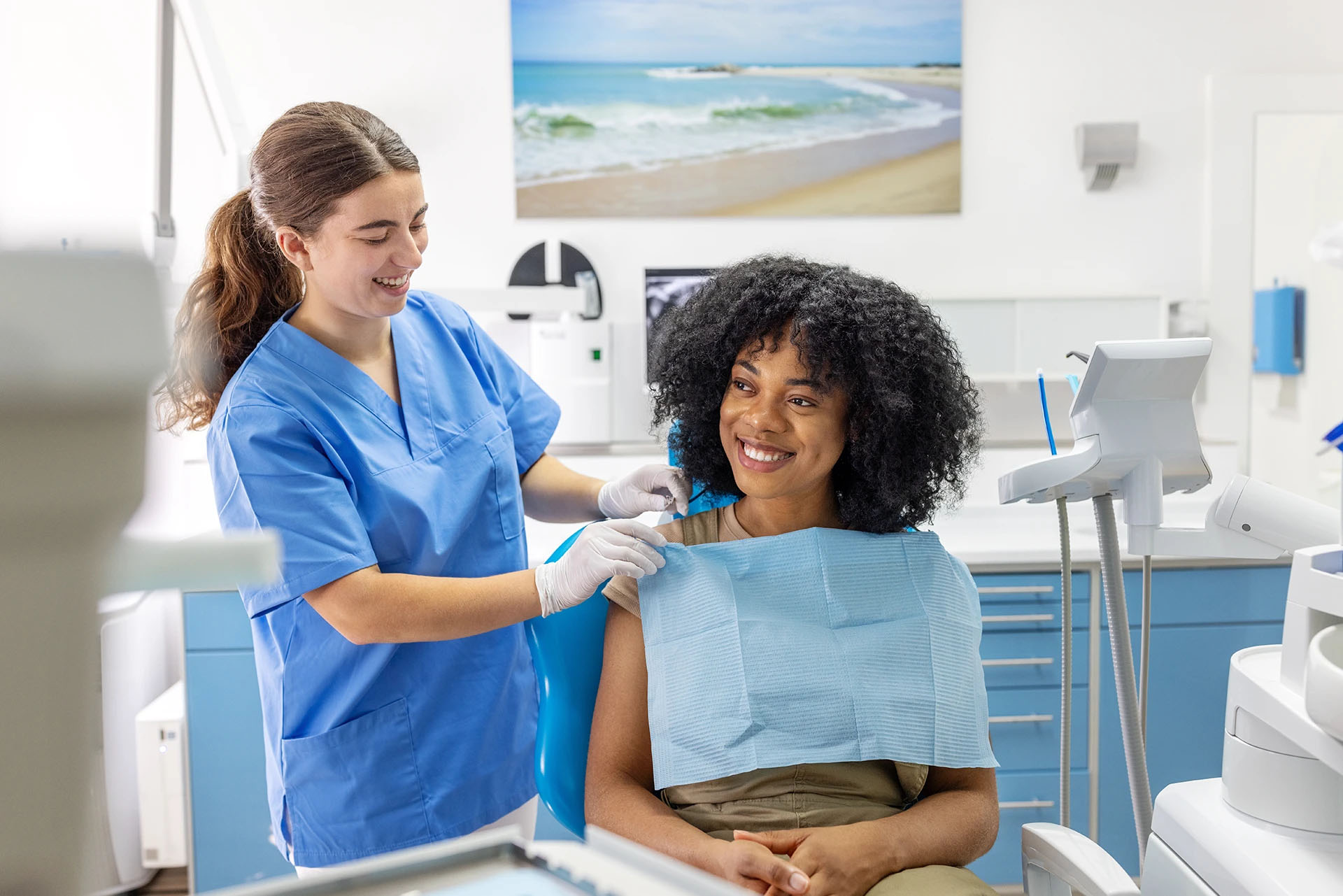 Dental hygienist in blue scrubs placing a bib on a smiling patient with a beach picture on the wall