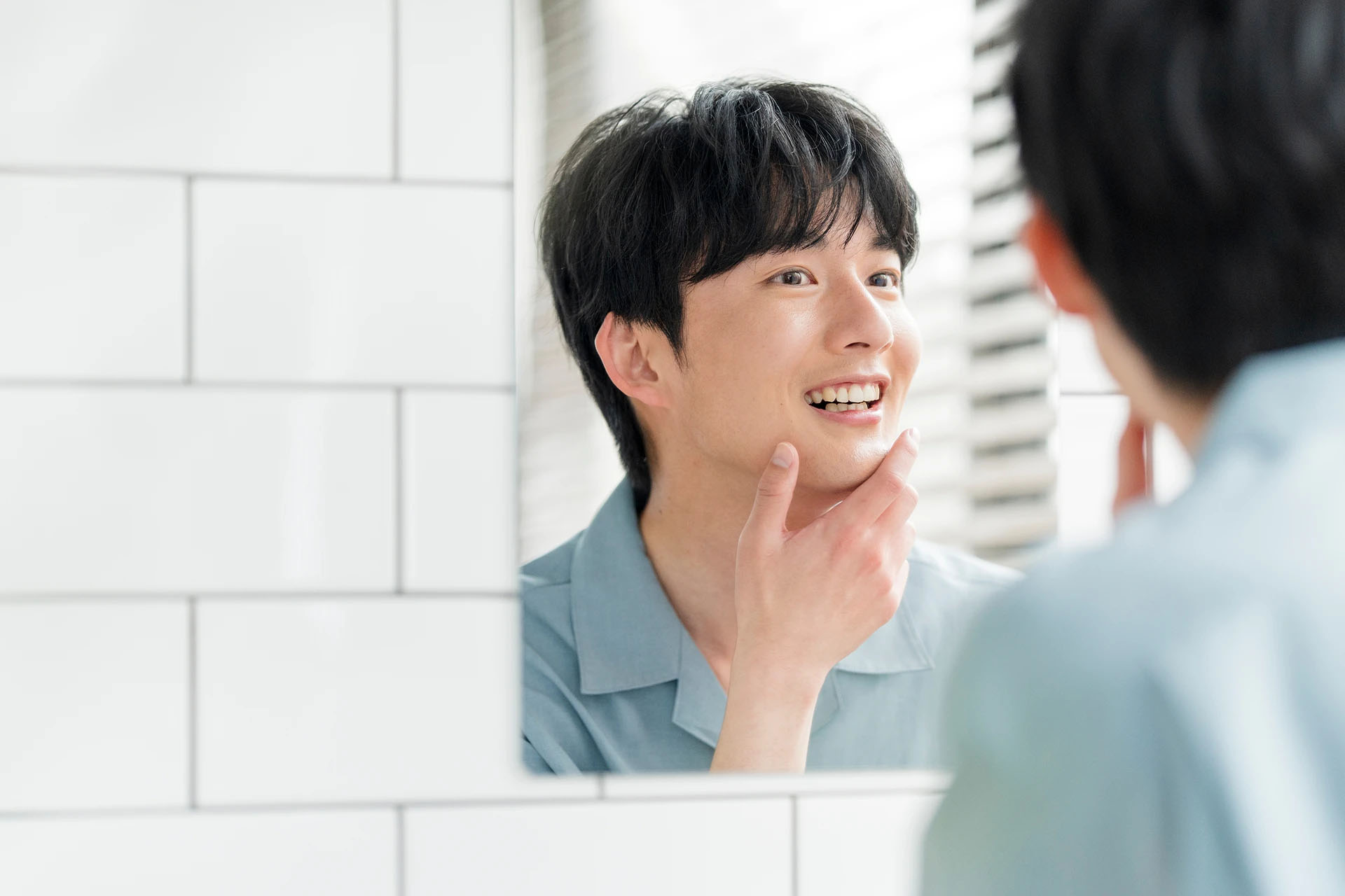 Smiling young man looking at his teeth in the bathroom mirror while touching his chin