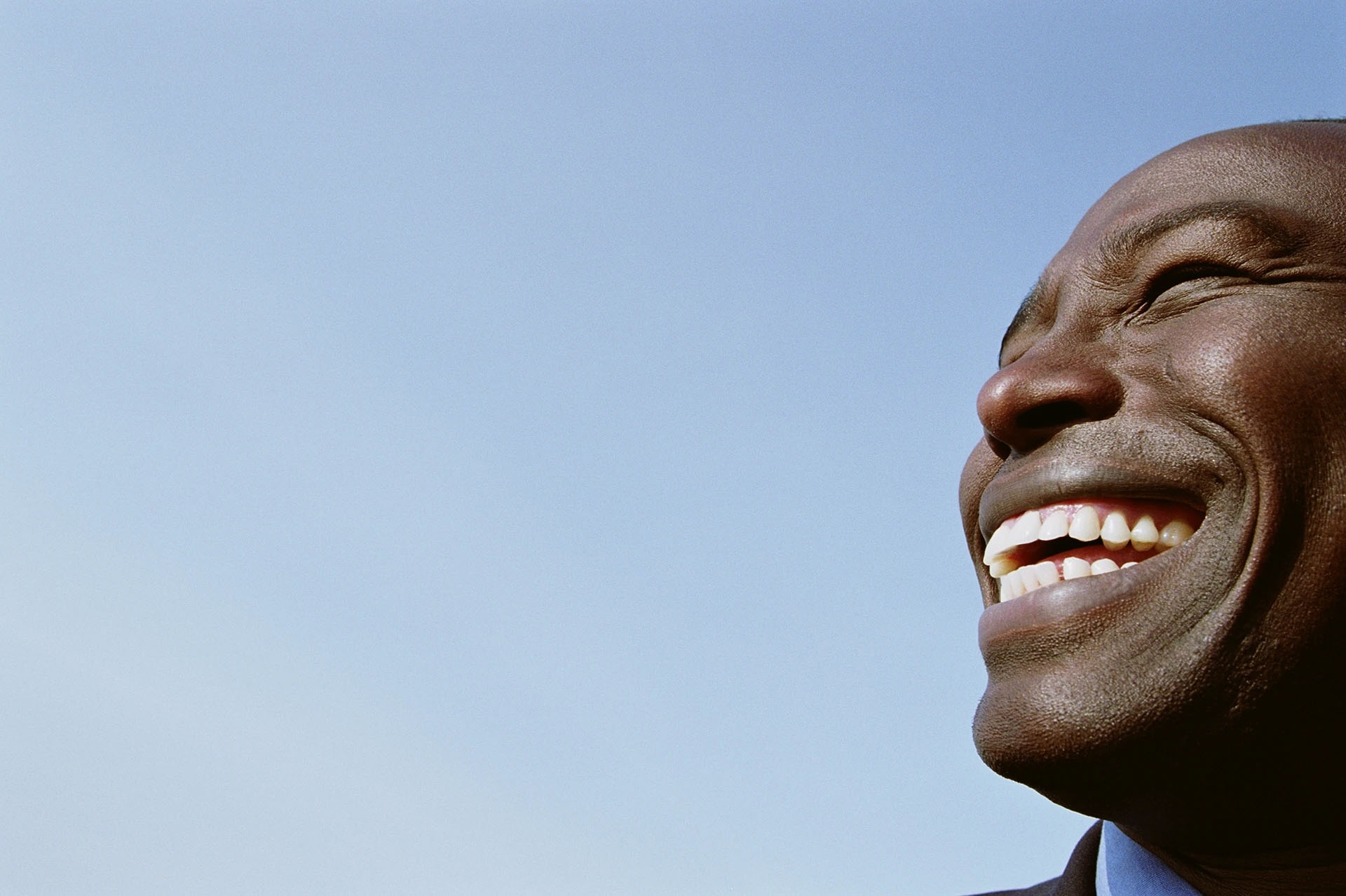 Close-up of a Black man laughing joyfully against a blue sky, showing a healthy white smile