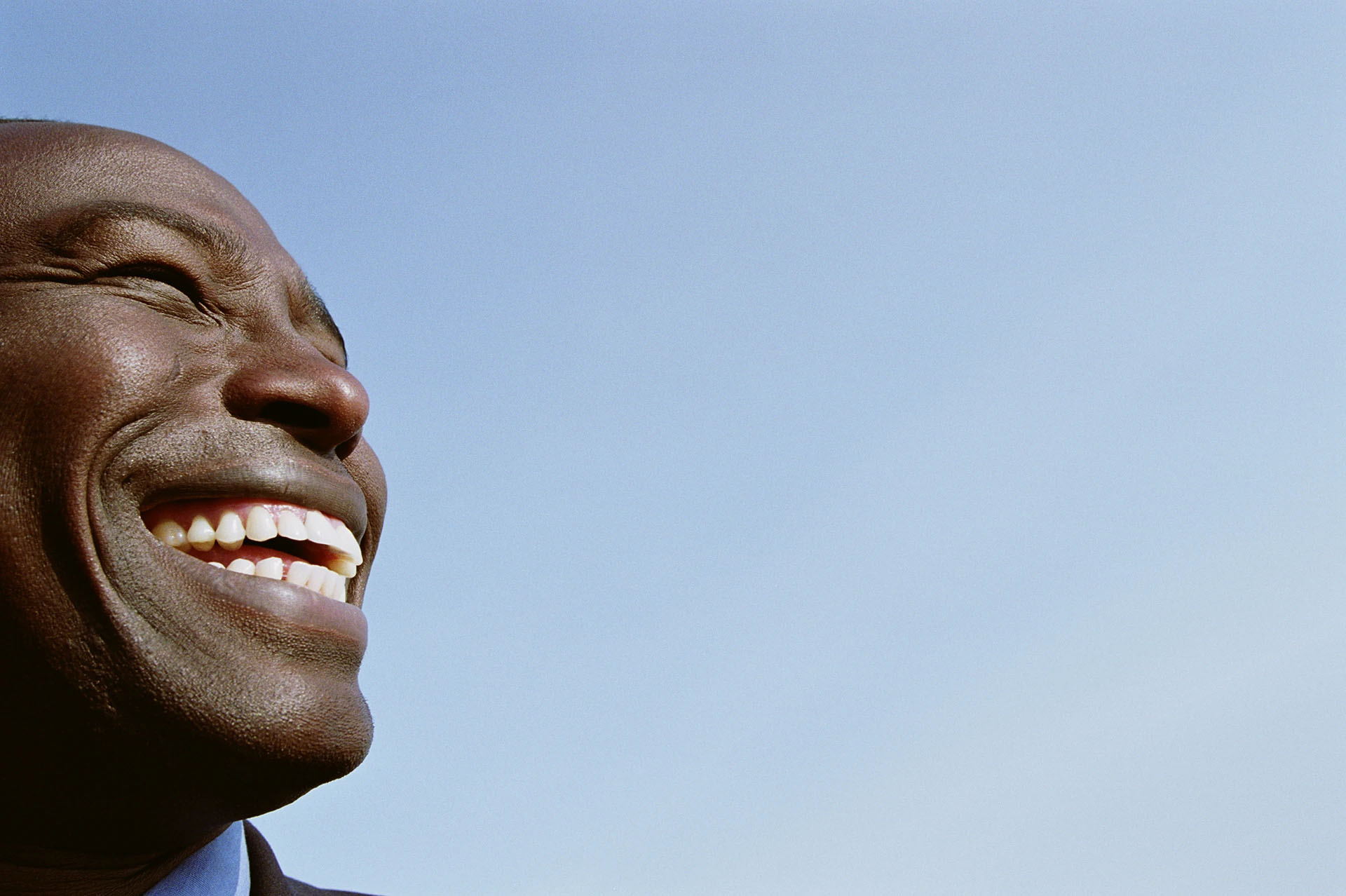 Close-up of a Black man laughing joyfully against a blue sky, showing a healthy white smile