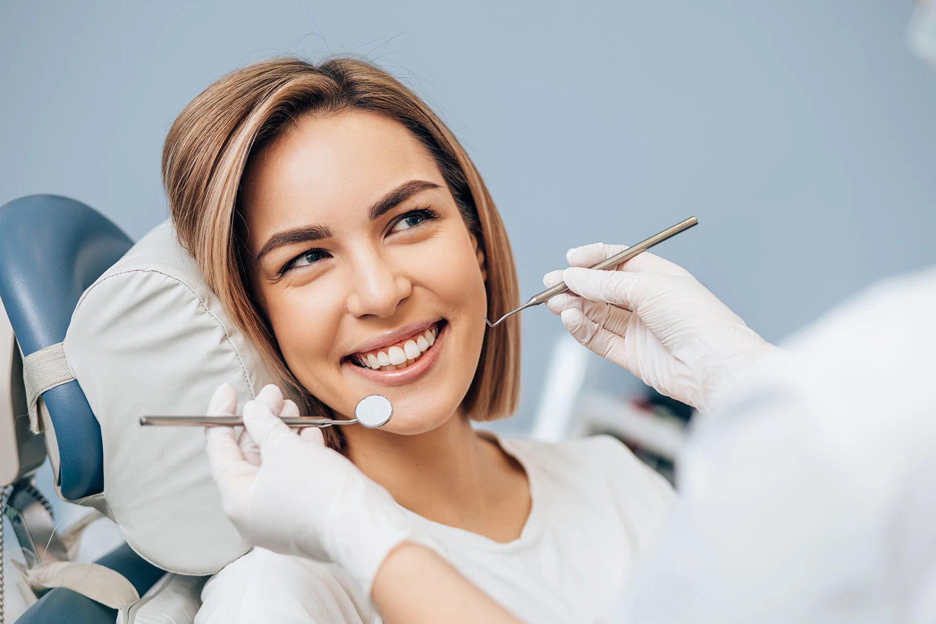 Smiling blonde woman in a dental chair against a blue background during a checkup with instruments nearby