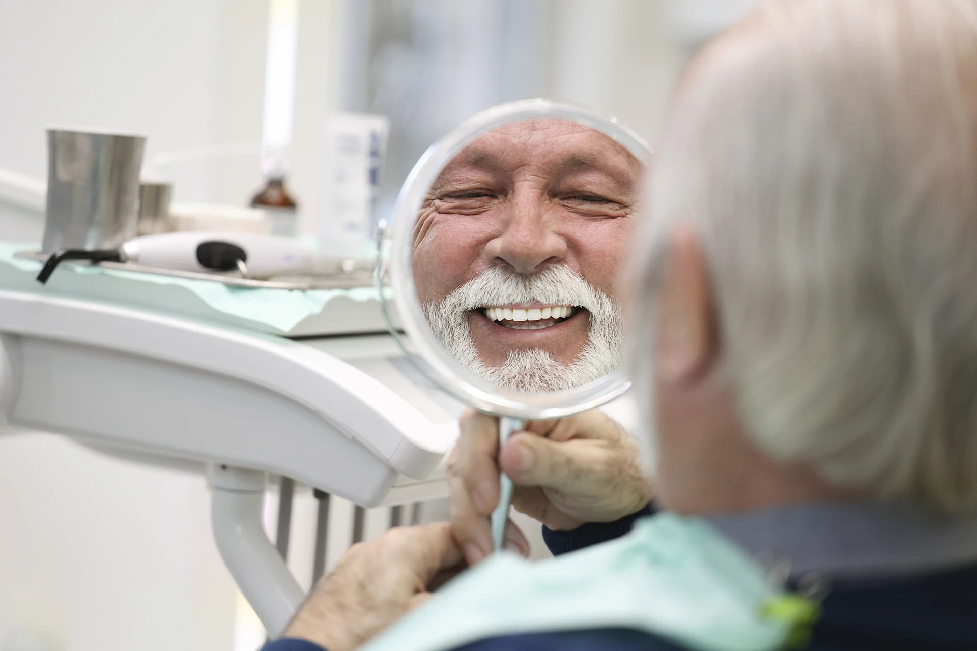 Smiling older man with a white beard looking at his teeth in a hand mirror in the dental office