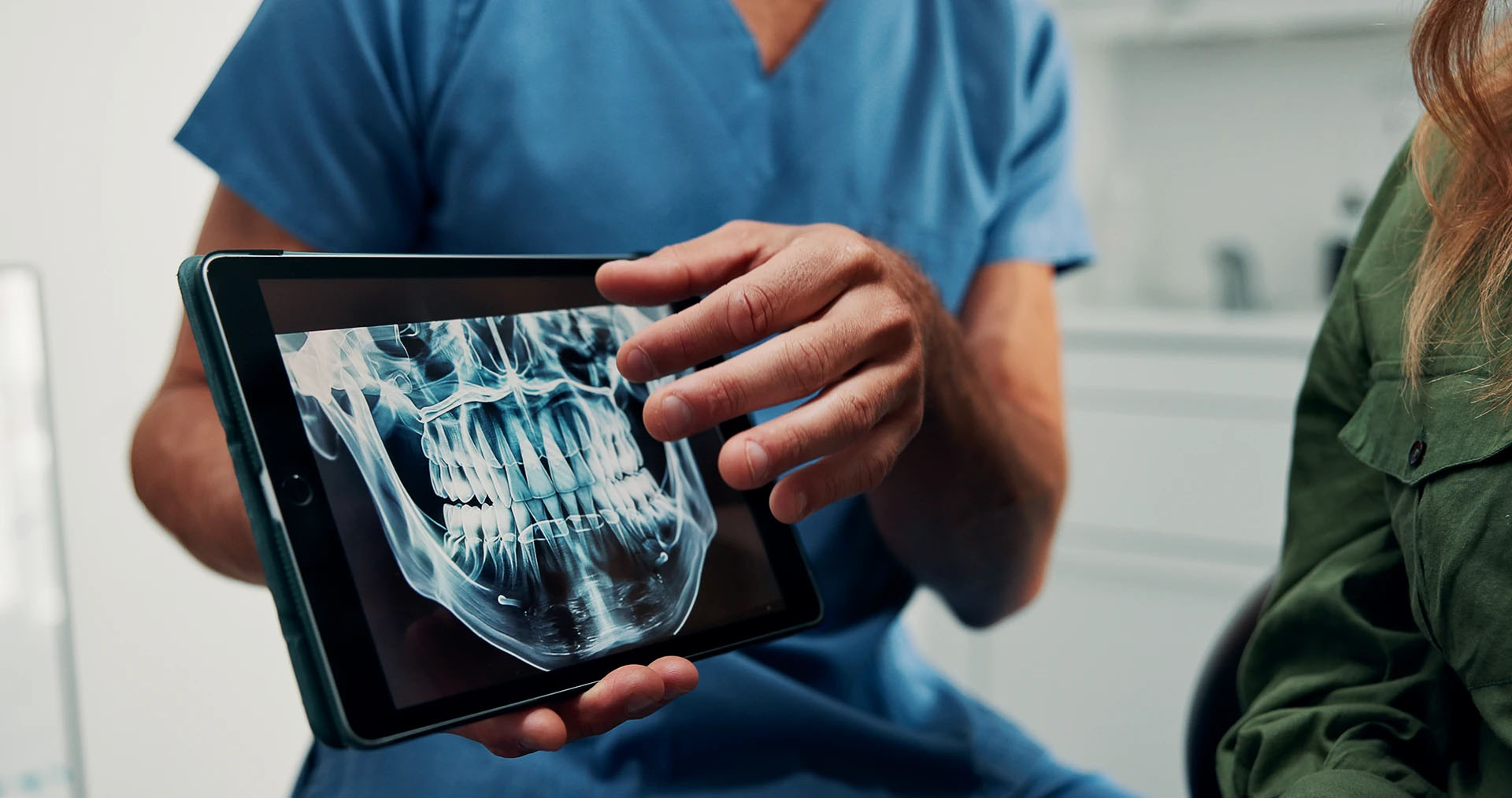 Dentist in blue scrubs holding a tablet displaying a panoramic dental X-ray beside a patient