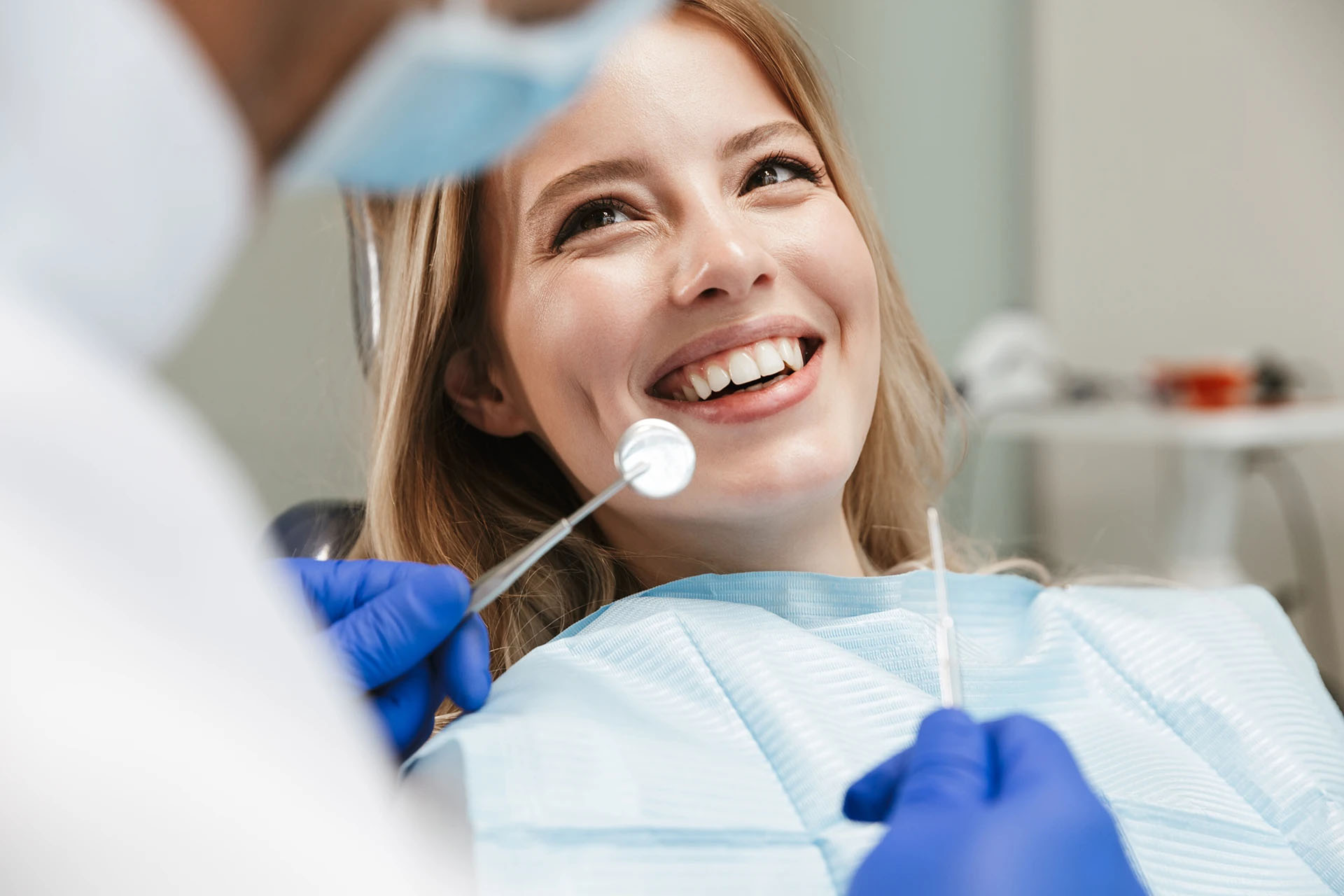 Blonde woman smiling in a dental chair as a dentist in blue gloves prepares a mirror and probe