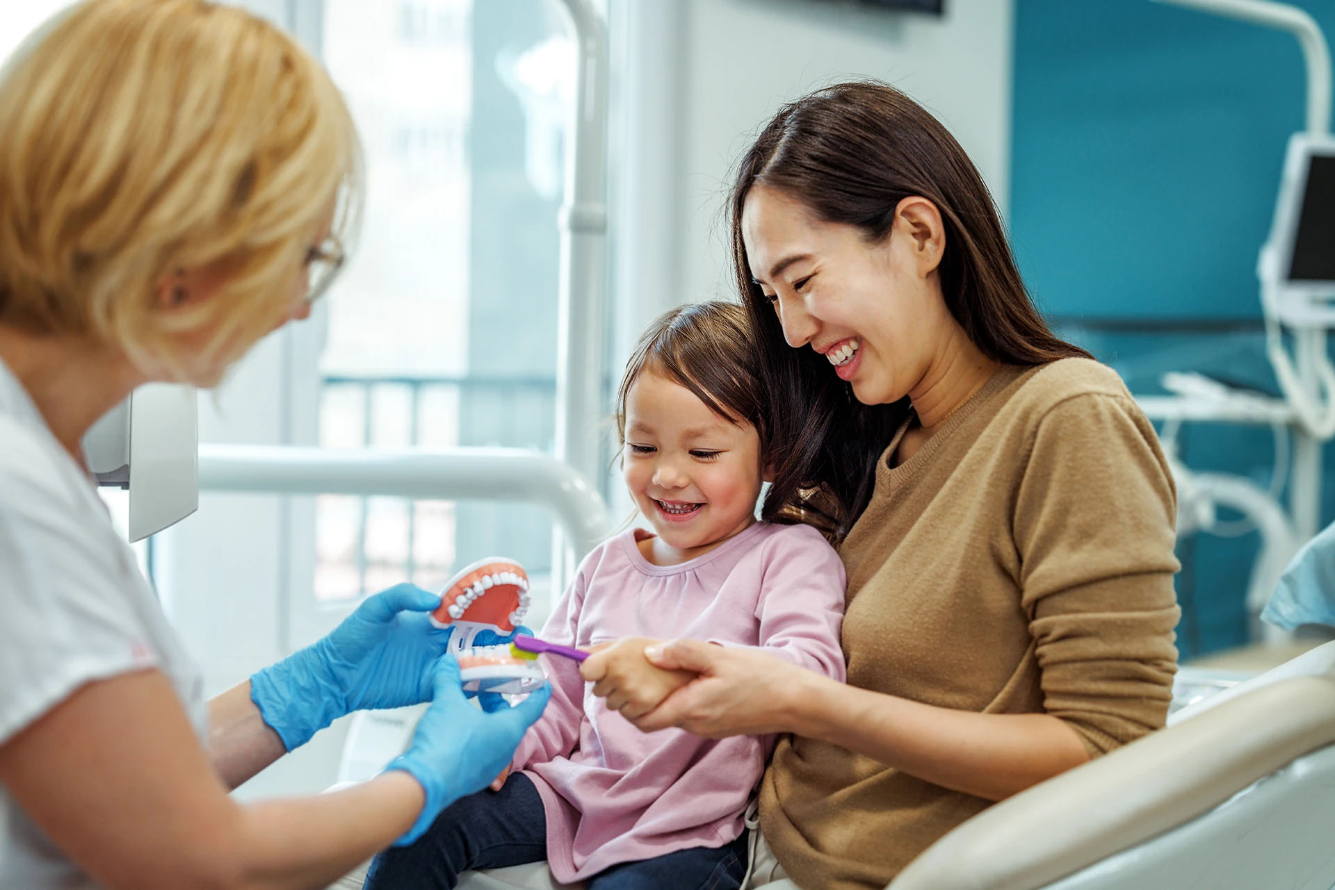 Dentist showing a little girl how to brush using a model while she sits on her smiling mother's lap