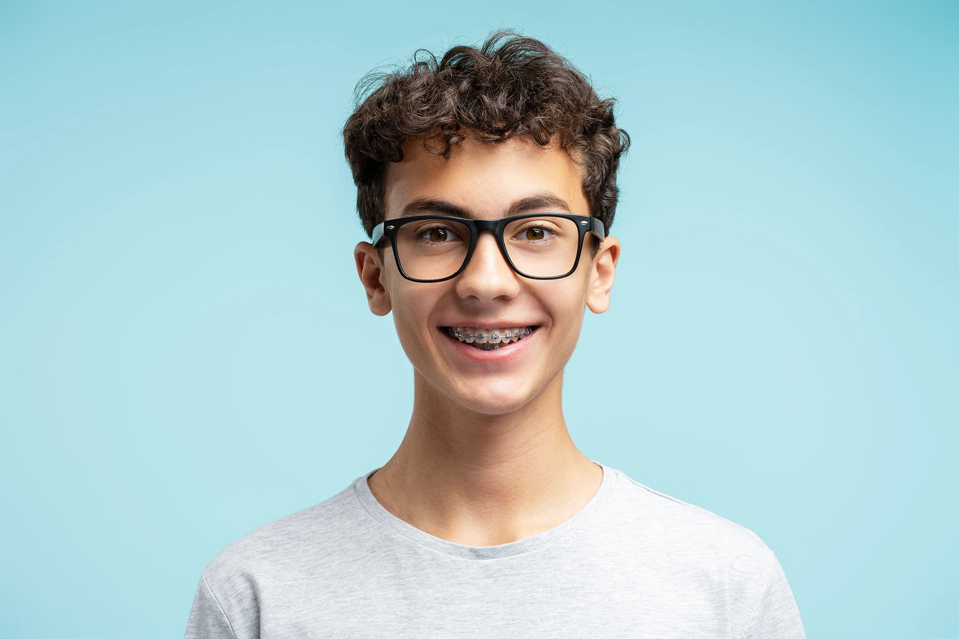Smiling teenage boy with glasses and metal braces against a blue background