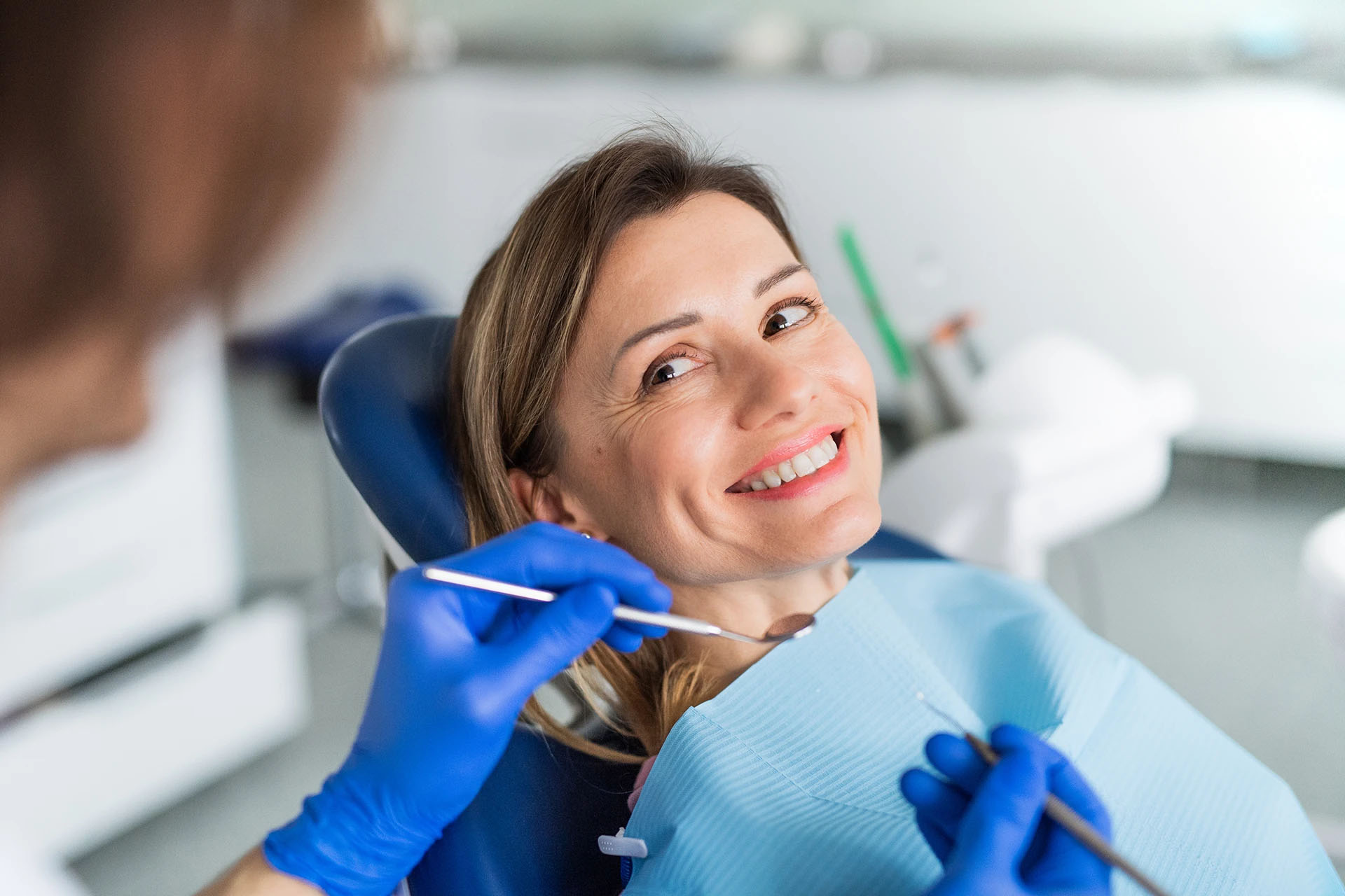 Woman reclined in a dental chair smiling while a dentist in blue gloves examines her teeth with a mirror