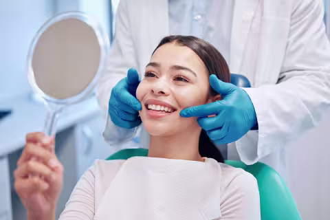 Smiling young woman in a dental chair looking in a hand mirror as a dentist in blue gloves frames her face