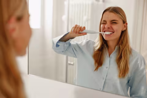 Blonde woman in a light blue shirt brushing her teeth and winking in the bathroom mirror